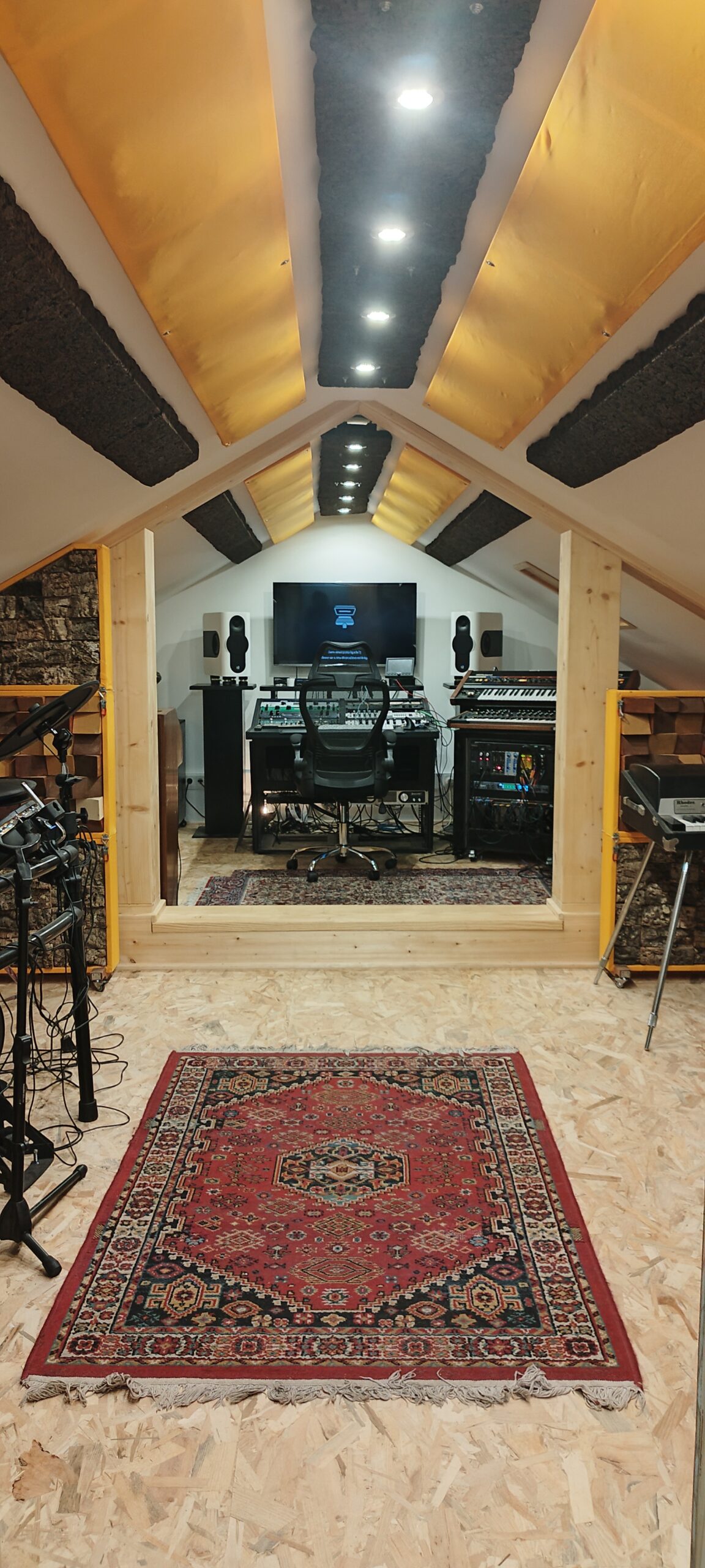 Recording equipment rests on a desk, flanked by speakers, with a large screen at the center, all within an attic setting.
